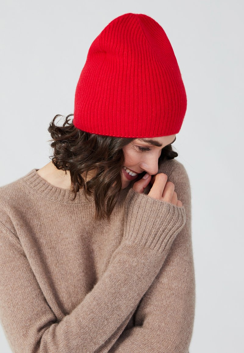 Rote, gerippte Strickmütze mit spitzem oberen Ende, getragen mit einem hellbraunen, weichen, langärmligen Pullover mit gerippten Bündchen.