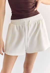 Pantaloni corti bianchi con elastico in vita e stringa di regolazione, caratterizzati da pieghe e una consistenza liscia. Abbinati a un top in cotone brown aderente.