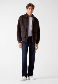 Veste zippée marron foncé avec deux grandes poches avant, portée sur une chemise blanche, associée à un jean bleu foncé et des baskets blanches.