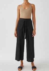 Pantalon large noir texturé avec une taille nouée, associé à un débardeur ajusté brun clair et des sandales à lanières tressées noires.