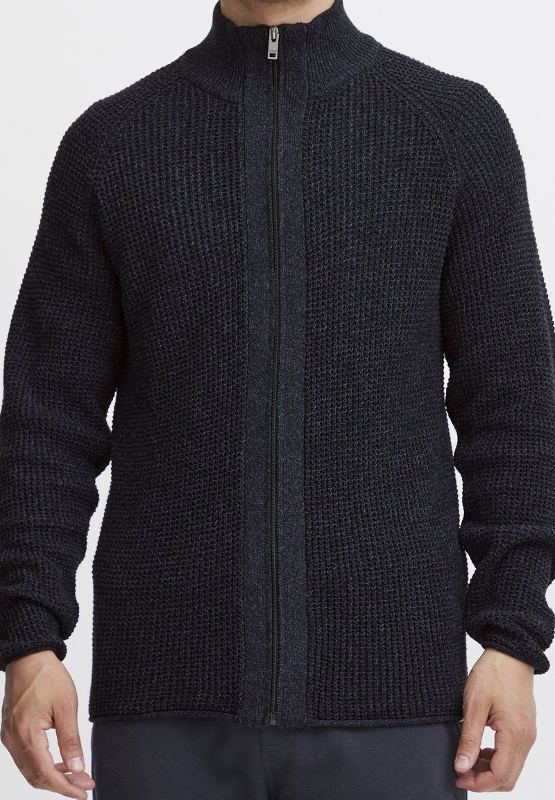 Homme portant un pull zippé noir en maille texturée avec col haut, debout devant un fond blanc uni.