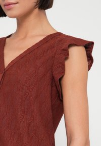 Μπλουζάκι σε χρώμα σκουριάς με υφή υφάσματος που παρουσιάζει κυματιστό μοτίβο, λαιμόκοψη V και ριχτά μανίκια βολάν. Λείο ύφασμα και καθημερινός σχεδιασμός.