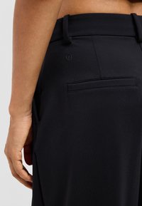 Close-up of a person’s lower back and hand wearing black pants with belt loops, a back pocket, and a small circular logo on the waistband.