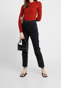 Rode geribbelde langetermijntop in combinatie met hoge zwarte jeans. Het model houdt een kleine zwarte beaded handtas vast en draagt zwarte gespen sandalen.