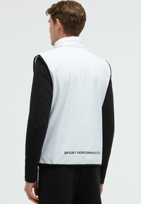 Chaleco sin mangas blanco con cuello alto, detalles negros en los bordes, un brillo sutil y el texto "SPORT PERFORMANCE ZERO" en el dobladillo.