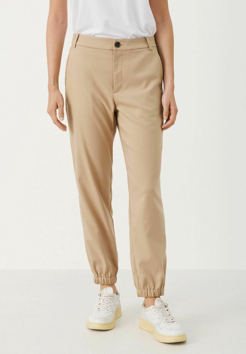 Beiga joggerbyxor i mjukt tyg med elastiska muddar, en framknäppning och sidofickor, tillsammans med vita sneakers.