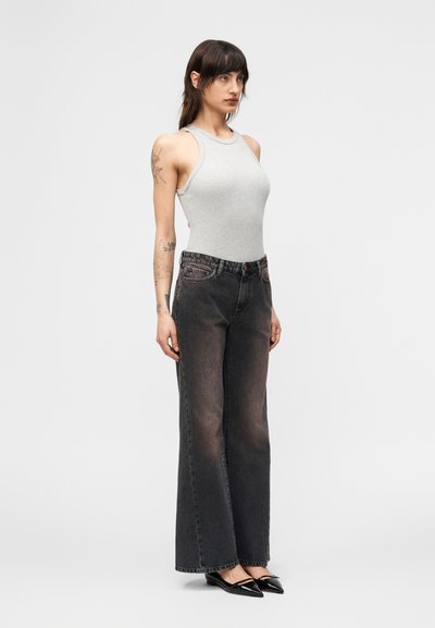 Γυναίκα με σκούρα μαλλιά μέχρι τους ώμους, φοράει γκρι αμάνικη μπλούζα, ξεβαμμένα μαύρα τζιν φαρδύ ποδόγυρο και μαύρα μυτερά φλατ παπούτσια, στέκεται πλαγιαστά.