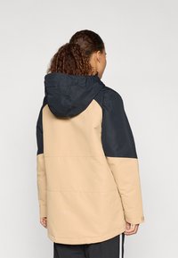 Vodotesný kabát v béžovej a námorníckej farbe, s kapucňou, kontrastnými panelmi a voľným strihom. Vyrobený z odolnej tkaniny s hladkou štruktúrou.