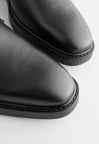 Fekete bőrcipő sima textúrával, hegyes orral és varrott fekete talppal. Minimalista díszítéssel és fényes felülettel rendelkezik.