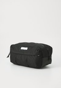 Schwarze Nylon-Aufbewahrungstasche mit Reißverschluss oben, gefalteter Textur, einem Markenetikett und einem kleinen Logo-Akzent an der Seite. Kompaktes Design.