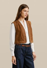 Hnedý suede vest s výstrihom do V, nosený cez bielu košeľu s gombíkmi. Skombinovaný s nohavicami z tmavého denimu s vysokým pásom a hnedým opaskom.