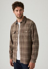 Uomo che indossa una camicia a quadri marrone sopra una t-shirt bianca e jeans scuri, in piedi contro uno sfondo grigio chiaro uniforme.