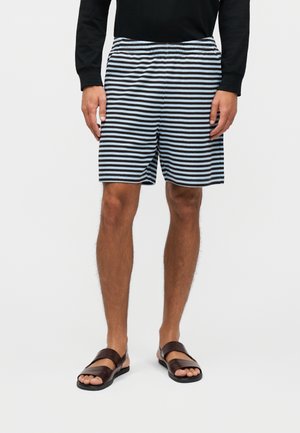 Hombre con pantalones cortos de rayas horizontales en negro y azul claro, camiseta negra de manga larga y sandalias marrón oscuro, de pie frente a un fondo liso.