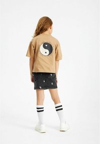 Tricou din bumbac bej cu un grafic yin-yang pe spate. Asociat cu o fustă din denim neagră cu modele repetate de yin-yang alb. Sosete albe cu dungi negre.