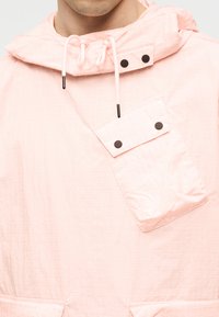 Ροζ ανοιχτό ροζ μπουφάν με κουκούλα, κορδόνια και μια πλάγια τσέπη στο στήθος που κλείνει με δύο μαύρα κουμπιά τύπου snap.