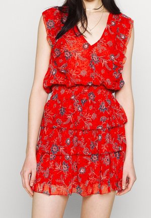 Robe sans manches rouge à motifs floraux avec des volants en couches, un décolleté en V et une taille ample, portée par une personne aux cheveux foncés et aux colliers superposés.