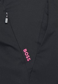 Svart tygjacka med kantig design, prydd med en rosa "BOSS"-logotyp och en liten dragkedjeficka på vänster sida. Slät textur.