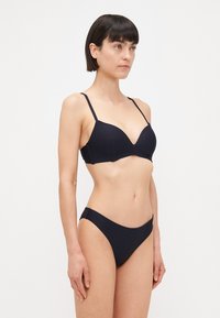 Crni bikini set s teksturiranim grudnjakom s dubokim izrezom, tankim podesivim trakama i odgovarajućim niskim gaćicama. Cjelokupna površina od glatkog materijala.