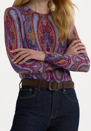 Femme aux longs cheveux blonds porte un pull coloré à motif cachemire, un jean foncé et une ceinture marron avec une boucle argentée, bras croisés.