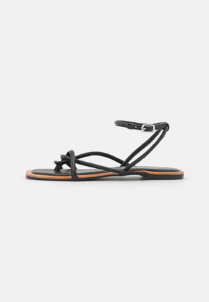 Sort flad sandal med tynde krydsede remme, ankelspænde og en tan kantssål på hvid baggrund.