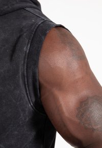 Mouwloze zwarte sweatshirt met geribbelde kraag; close-up van een gespierde arm met tatoeages en een gladde huidskleur. Textuur van de stof zichtbaar.