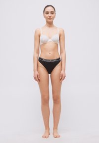 Γυναίκα που στέκεται ξυπόλυτη με το πρόσωπο προς τα εμπρός, φορώντας λευκό δαντελωτό σουτιέν και μαύρα εσώρουχα Polo Ralph Lauren, σε απλό ανοιχτό φόντο.