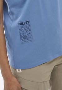 Niebieska bawełniana koszulka z grafiką przedstawiającą kwiatowe wzory oraz logo "Millet"; zestawiona z beżowymi spodniami. Miękka tekstura, casualowy krój.