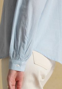 Light blue, long-sleeve shirt with thin vertical stripes worn with off-white pants, showing cuff with button and part of back pocket.