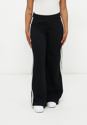 Person wearing black wide-leg pants with white side stripes and white sneakers, standing against a plain white background.