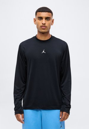 Muž s krátkymi vlasmi, ktorý má na sebe čierne tričko s dlhým rukávom a modré nohavice, oboje s malým bielym logom Jumpman.