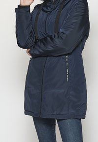 Chaqueta acolchada azul marino con capucha, con cordones negros, cremallera frontal y dos bolsillos laterales con cremallera. Textura lisa y brillante.