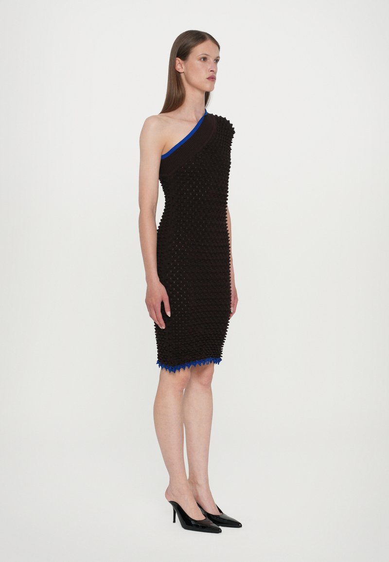 Asymmetrisk brun kjole med tekstureret detaljering, blå udskæring og spidse kanter. Sammen med sorte højhælede mule-sko.