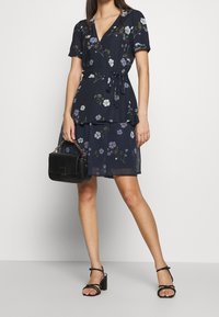 Robe portefeuille marine à imprimé floral avec manches courtes, taille nouée, jupe à volants et sac à main noir. Des talons noirs à brides complètent la tenue.