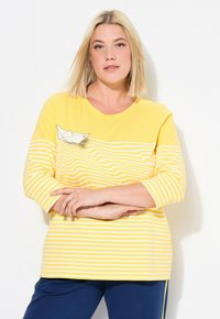 Gul långärmad skjorta med vita horisontella ränder, med en dekorativ pappersbåt-design. Mjuk bomullstyg med sydda detaljer.