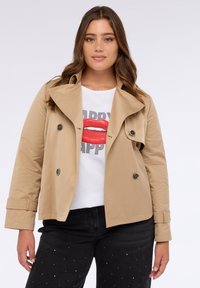 Ljusbeige trenchcoat med spetsig krage och dubbelknäppt design. Har svarta knappar och en mjuk, slät tygtextur.