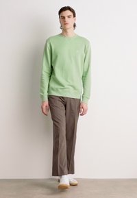 Groene crewneck sweatshirt gemaakt van zacht materiaal, gecombineerd met bruin op maat gemaakte broek. Model draagt witte sneakers. Eenvoudig, modern ontwerp.