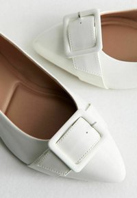 Vita läderskor med platt klack och spetsig tå, med en stor fyrkantig spänne detaljer samt en mjuk brun inredning.