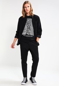 Sort fløjlsskjorte over en sort T-shirt med hvid grafisk design. Et par sorte bukser og sorte sneakers med hvide såler.