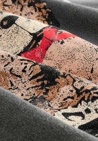 Gros plan sur un tissu texturé avec des motifs abstraits en noir, marron et rouge sur un fond beige, encadré de plis gris doux.