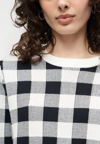 Γυναίκα με καστανά σγουρά μαλλιά, φορεί κρίκους σκουλαρίκια και ασπρόμαυρο πουλόβερ με καρό μοτίβο και στρογγυλή λαιμόκοψη.
