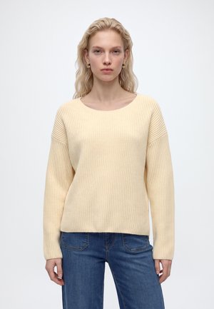 Blonde vrouw met golvend haar, die een crèmegekleurde gebreide trui en een blauwe spijkerbroek draagt, staand tegen een effen witte achtergrond.