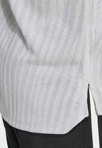 Svetlo šedá košeľa s textúrovaným vzorom vertikálnych línií a malých dierok. Obsahuje bočné rozparky a hladký povrch látky.