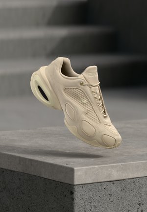 Beżowy but sportowy z teksturowaną cholewką i unikalną podeszwą, unoszący się nad szarą betonową krawędzią z rozmytymi schodami w tle.