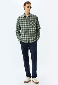 Grön- och marinblå rutigt flanellskjorta med bröstficka, bärs över en vit t-shirt. Marinblå byxor och ljusa sneakers fulländar looken.