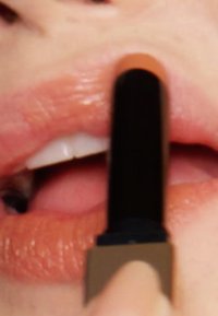Un tube de rouge à lèvres tenu près des lèvres ; le tube a une forme cylindrique noire, avec une finition mate, tandis que les lèvres sont de couleur pêche et brillantes.