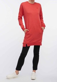 Robe sweat rouge avec des manches longues, un col rond, des poches latérales et des fentes sur le bas, associée à un pantalon noir et des baskets blanches.
