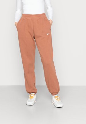 Personne portant un pantalon de survêtement Nike couleur rouille avec des poignets élastiques et des baskets blanches avec des lacets orange, debout devant un fond uni.
