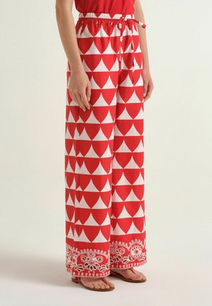 Femme portant un pantalon large rouge et blanc avec des motifs géométriques et floraux, accompagné de sandales plates et d'un haut rouge.