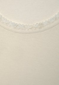 Cremfarbener, gerippter Stoff mit einer gewellten Spitzenborte entlang des Ausschnitts, der eine weiche Textur und eine körpernahe Passform zeigt.