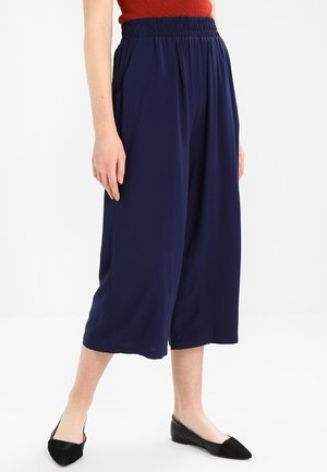 Mujer con pantalones anchos de pierna corta azul marino con cintura elástica y bailarinas negras puntiagudas sobre un fondo blanco liso.